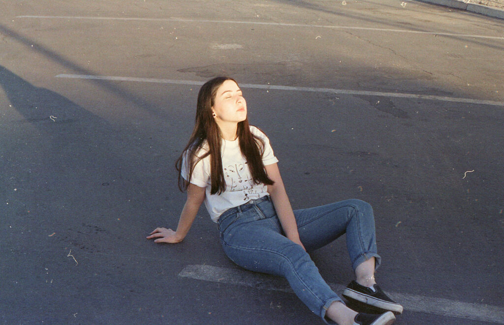 Film Photo of Gabriela Inzunza a 21-year-old from Los Angeles CA sitting on a parking lot during sunset. She wears a white t-shirt with a graphic design blue jeans and black and white sneakers.